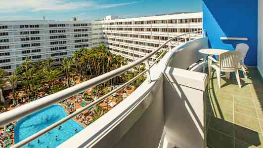 Udsigt fra en balkon mod et poolområde med mange liggestole og grønne områder, omgivet af hvide hotelbygninger. (AI-genereret tekst)