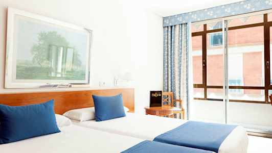 Et hotelværelse med to enkeltsenge, der har blå tæpper og puder, en stol ved et bord samt et vindue med blå gardiner. (AI-genereret tekst)