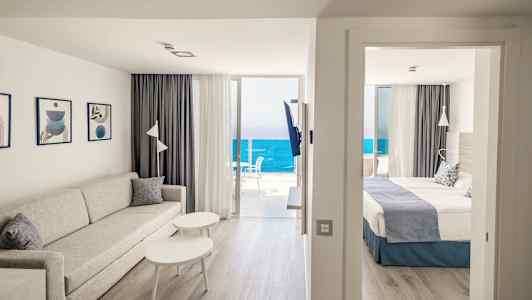 Stue med sofa og bord ved siden af soveværelset med dobbeltseng. Havudsigt fra altanen. (AI-genereret tekst)