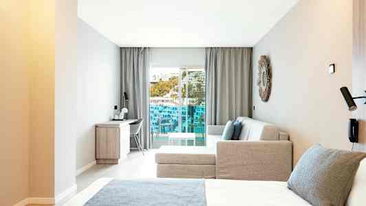 Moderne hotelværelse med seng, sofa og skrivebord samt en altan med udsigt. (AI-genereret tekst)