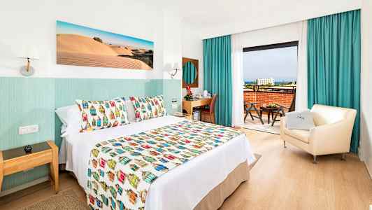 Hotelværelse med dekorativt sengetæppe, lænestol og skrivebord samt udsigt fra altanen. (AI-genereret tekst)