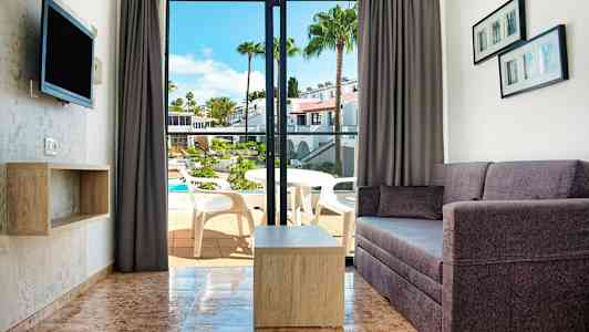 Udsigt over en stue med sofa og bord samt en åbning til en balkon med udsigt over en pool. (AI-genereret tekst)