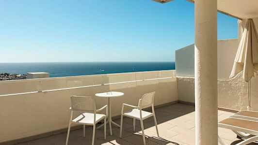 Terrasse med to hvide stole og et bord, havet ses i baggrunden. (AI-genereret tekst)