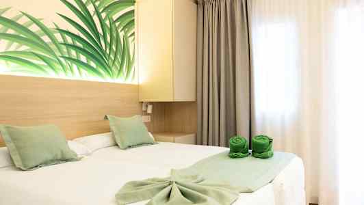 Et hotelværelse med to enkeltsenge med hvide lagner og grønne dekorative puder. Vægdekoration med bladmotiv samt grønne sammenrullede håndklæder på sengen. (AI-genereret tekst)