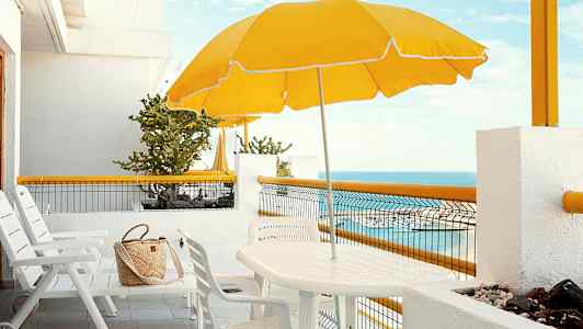 Hvid terrasse med stole, bord, gul parasol og havudsigt. (AI-genereret tekst)