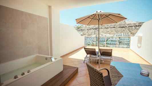 Terrasse med boblebad, parasol, liggestole og udsigt til huse på en bjergskråning. (AI-genereret tekst)