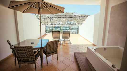 Terrasse med spisebord, parasol, liggestole og jacuzzi, med udsigt over omgivelserne. (AI-genereret tekst)