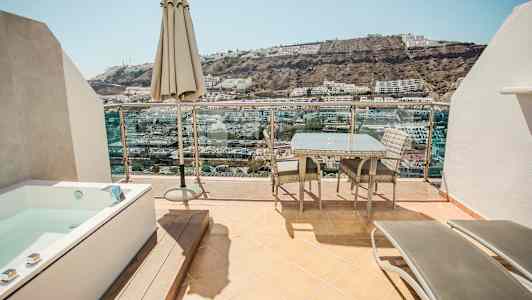 En terrasse med havemøbler, en parasol og et boblebad med udsigt over en skråning med bygninger. (AI-genereret tekst)