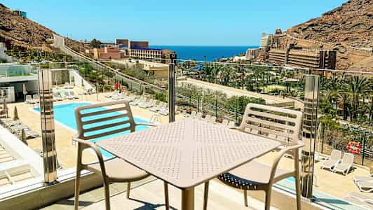 Udsigt over en pool og solsenge med havet og bygninger i baggrunden, set fra en balkon med bord og stole. (AI-genereret tekst)