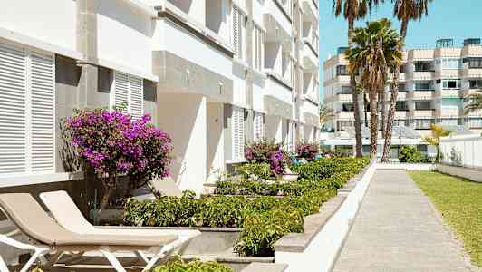 Billede af en hvid bygning med lilla blomster og liggestole ved en gangsti. (AI-genereret tekst)