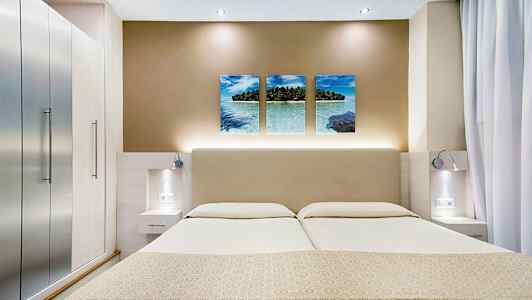 Billedet viser et moderne hotelværelse med en dobbeltseng, stilfulde sideborde og vægdekoration med tre billeder af et tropisk strandmotiv. (AI-genereret tekst)