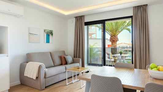 En stue med en grå sofa, et sofabord og et spisebord. Udenfor ses en terrasse med havemøbler og en palme. (AI-genereret tekst)