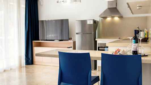 Et køkken med en køkkenbord, to blå stole, et køleskab og et fjernsyn ved et træskab. (AI-genereret tekst)