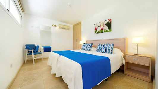 Et lyst hotelværelse med en dobbeltseng, blå og hvide sengelinned, natbord, vægdekoration og en lænestol. (AI-genereret tekst)