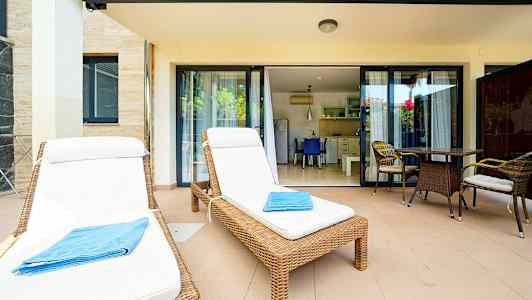 To liggestole med hvide hynder og et blåt håndklæde på en terrasse med bord og stole foran glasdøre til et køkken. (AI-genereret tekst)