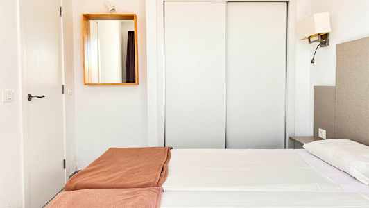 Et hotelværelse med hvidt interiør, en dobbeltseng med hvide lagner og brune sengetæpper samt et spejl på væggen. (AI-genereret tekst)