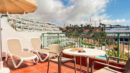 En terrasse med to solsenge, et bord og stole samt udsigt over et poolområde og palmer. (AI-genereret tekst)