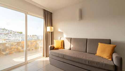 En grå sofa med gule puder står i et rum med store glasdøre, der fører ud til en altan. (AI-genereret tekst)