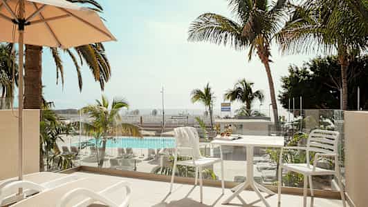 En altan med liggestole og et bord med stole, udsigt til en pool og palmer. (AI-genereret tekst)