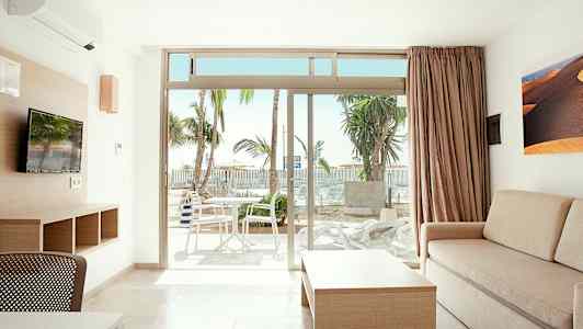 Et lyst rum med sofa, bord og TV samt udsigt til en terrasse med bord og stole. Palme træer kan ses udenfor. (AI-genereret tekst)