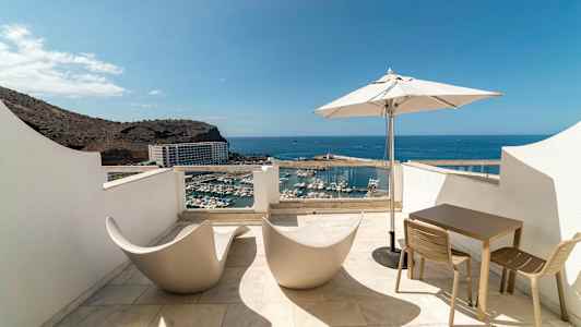 Terrasse med udsigt over havn og hav, udstyret med moderne stole, bord og parasol. (AI-genereret tekst)