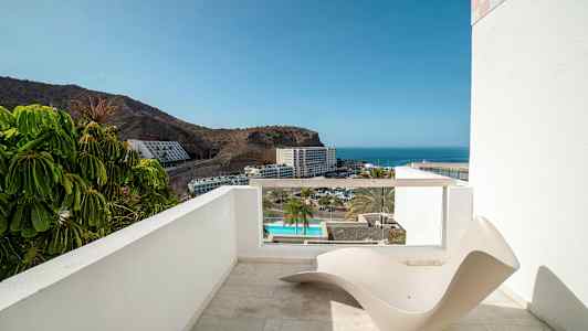 En terrasse med udsigt over havet og et hotelområde, med en minimalistisk liggestol i forgrunden. (AI-genereret tekst)