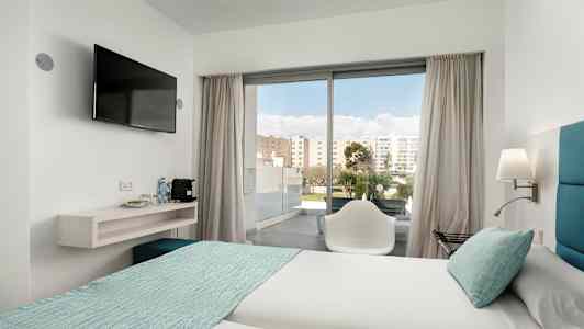 Et hotelværelse med to senge redt med lyse lagener og turkis sengetæppe. En stol står foran en glasdør, der fører ud til en altan. Et vægmonteret TV hænger ved siden af en hylde med vandflasker og kaffemaskine. (AI-genereret tekst)