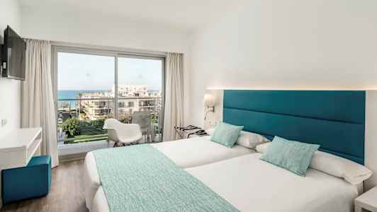 Et lyst hotelværelse med to enkeltsenge, store vinduer og udsigt over havet. (AI-genereret tekst)