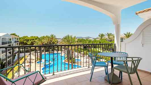 Udsigt fra en altan med bord og stole mod en pool og omgivelser med grønne områder. (AI-genereret tekst)