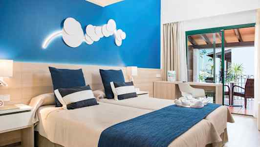 Et hotelværelse med to enkeltsenge, en blå væg, et skrivebord og en møbleret altan. (AI-genereret tekst)