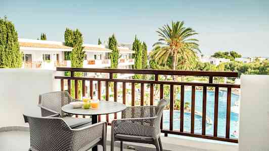 Udsigt fra en balkon med et bord og stole, hvor morgenmad er fremstillet. I baggrunden er der en pool, grøntområder og bygninger. (AI-genereret tekst)