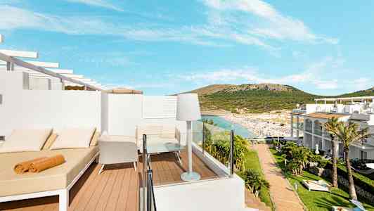 Udsigt over en terrasse med solsenge og stole, med sandstrand og omkringliggende bakker i baggrunden. (AI-genereret tekst)