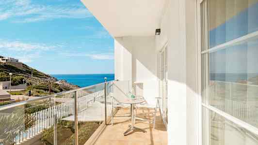 En altan med glasrækværk, et bord og to stole, med udsigt mod havet og det omkringliggende landskab. (AI-genereret tekst)