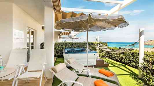 Terrasse med liggestole, parasol, jacuzzi og hængekøje samt udsigt over havet. (AI-genereret tekst)