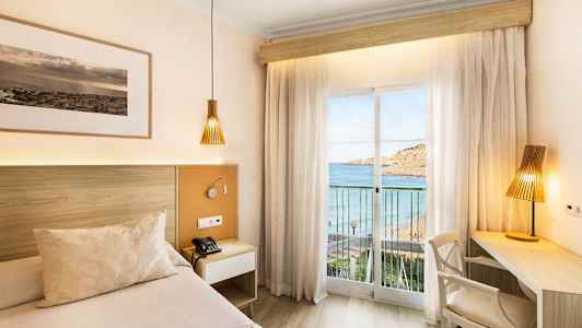 Et hotelværelse med en seng og skrivebord, med udsigt til havet gennem et glasvindue med gardiner. (AI-genereret tekst)