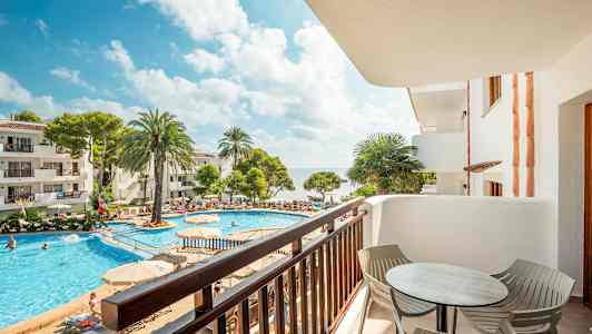 Udsigt fra en altan over en pool omgivet af liggestole, palmer og bygninger. (AI-genereret tekst)