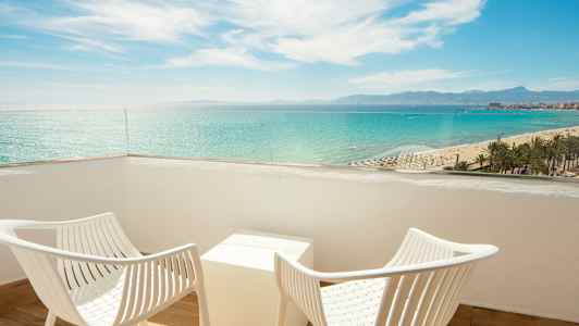 To hvide stole og et bord på en balkon med udsigt over havet og stranden. (AI-genereret tekst)