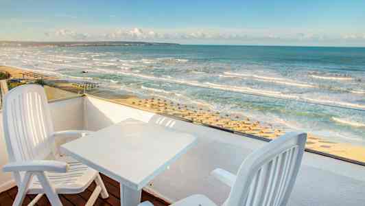 Udsigt fra en altan med to stole og et bord mod en strand med parasoller og bølgende hav. (AI-genereret tekst)