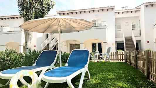 To solsenge med en parasol på en græsplæne foran en hvid bygning. (AI-genereret tekst)