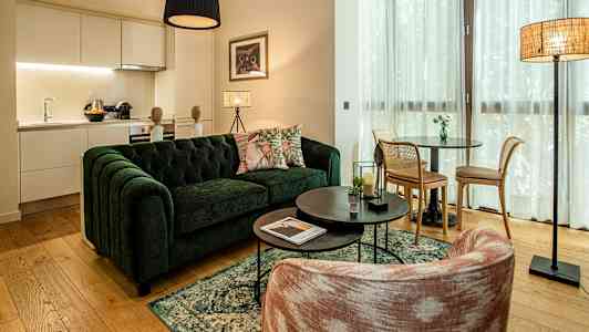 En stue med en grøn sofa, et sofabord og en spiseplads. Køkkenområdet er synligt i baggrunden. (AI-genereret tekst)