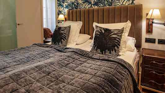 Et soveværelse med en seng med brune puder, gråt sengetæppe og en mønstret væg bagved. (AI-genereret tekst)