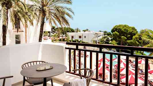 Udsigt fra en balkon med stole og bord mod en pool og omkringliggende haver. (AI-genereret tekst)