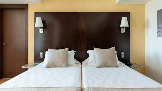 To enkeltsenge med hvide lagner og beige puder mod en mørkebrun sengegavl, med væglamper over. (AI-genereret tekst)