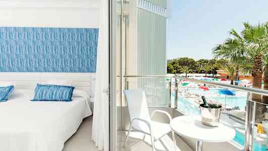 Et hotelværelse med to senge, et blåt sengegærde og adgang til en balkon med udsigt over en pool og have. (AI-genereret tekst)