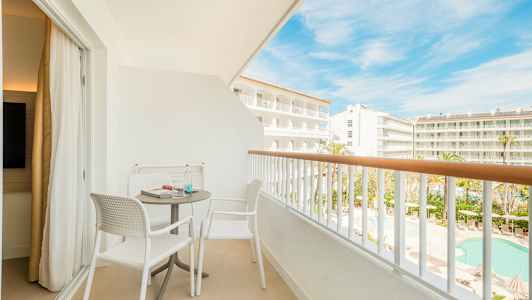 En møbleret altan med to stole og et bord, med udsigt over hotelområdet og poolen. (AI-genereret tekst)