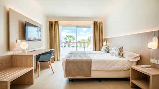 Moderne indrettet hotelværelse med seng, skrivebord og store glasdøre ud til en balkon. (AI-genereret tekst)