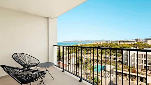 Altan med to sorte stole og bord, udsigt over hotelområde og hav. (AI-genereret tekst)