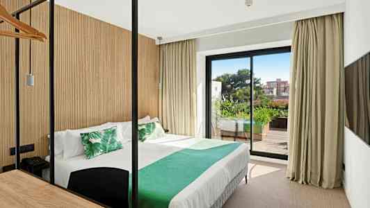 Et hotelværelse med en seng redt med hvide lagner og grønne puder, en stor glasdør der fører ud til en altan med beplantning. (AI-genereret tekst)