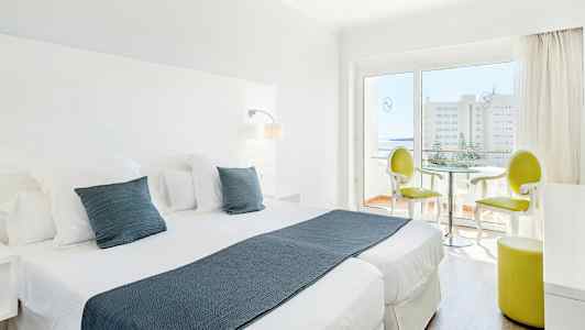 Et lyst hotelværelse med to enkeltsenge, hvide møbler og en siddegruppe nær en glasdør, der fører til en balkon. (AI-genereret tekst)