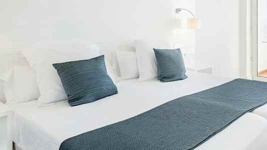 Hvide sengelinned på en dobbeltseng med blå pyntepuder og sengetæppe. (AI-genereret tekst)